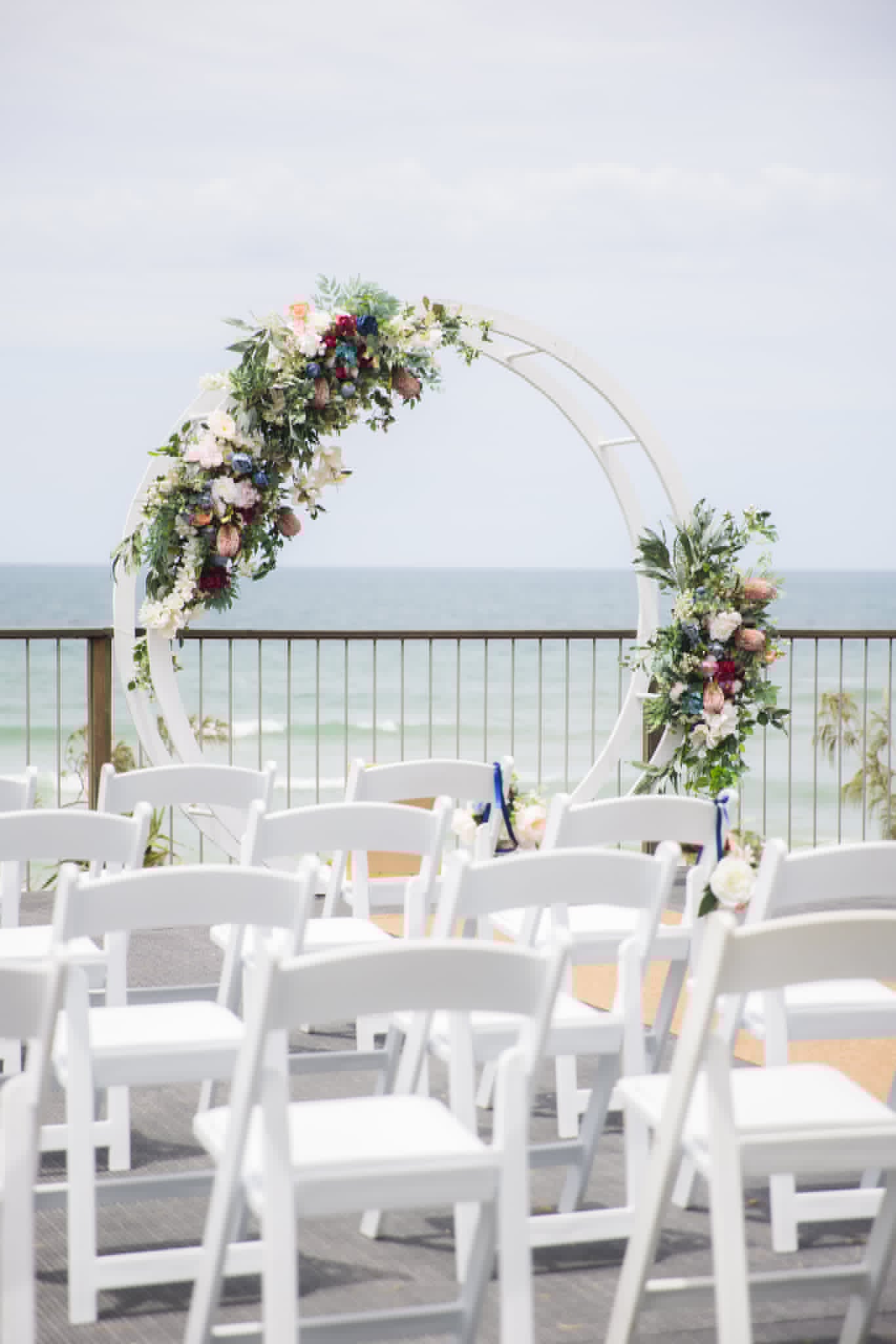 white chairs in front of a white arch with leaves and a water and sky background
